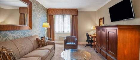 A hotel room with a sofa, a glass coffee table, a leather armchair, a wooden cabinet, a flat-screen TV, and a window with curtains.