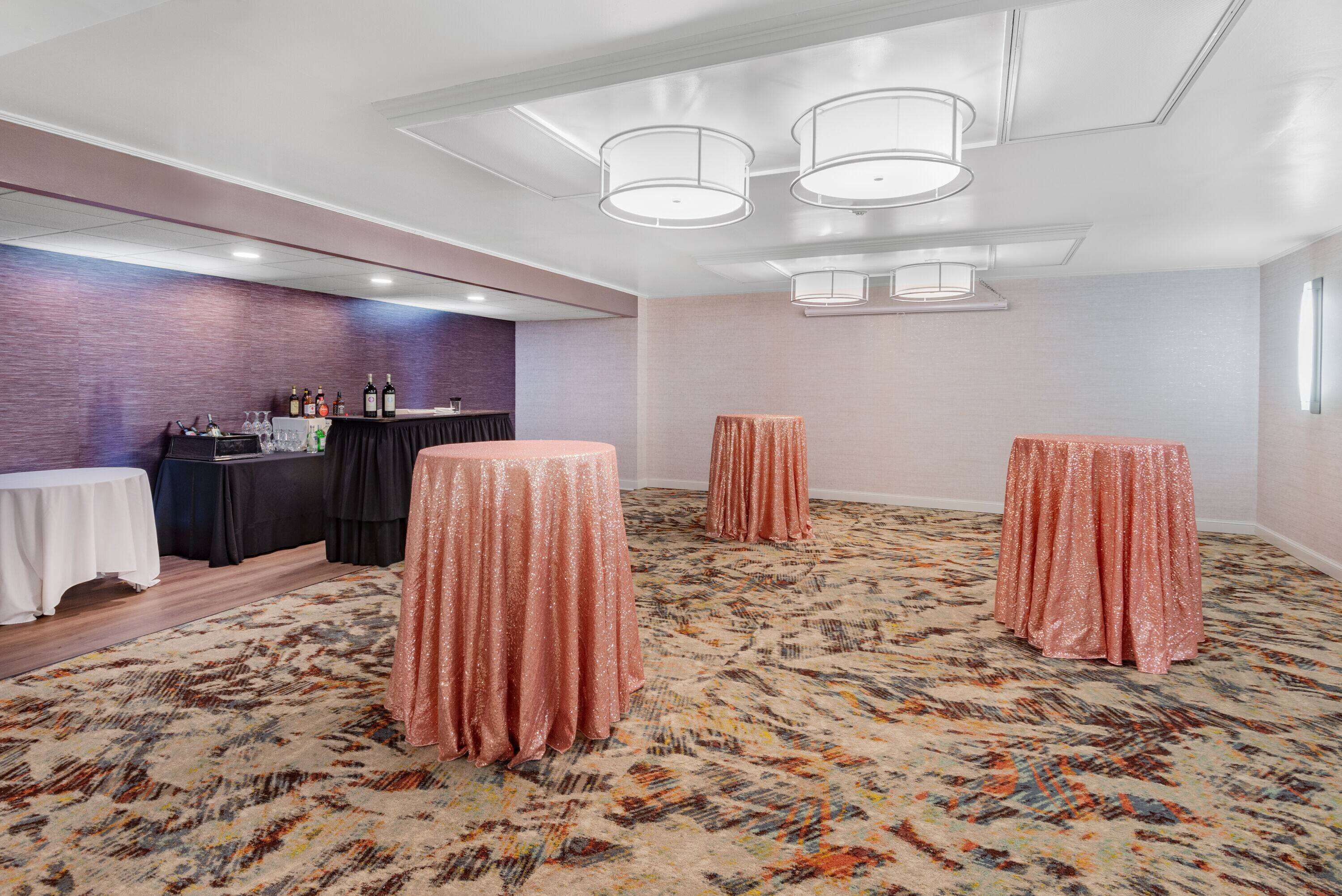 A conference room with a patterned carpet, a bar area with bottles, and three round tables with draped covers.