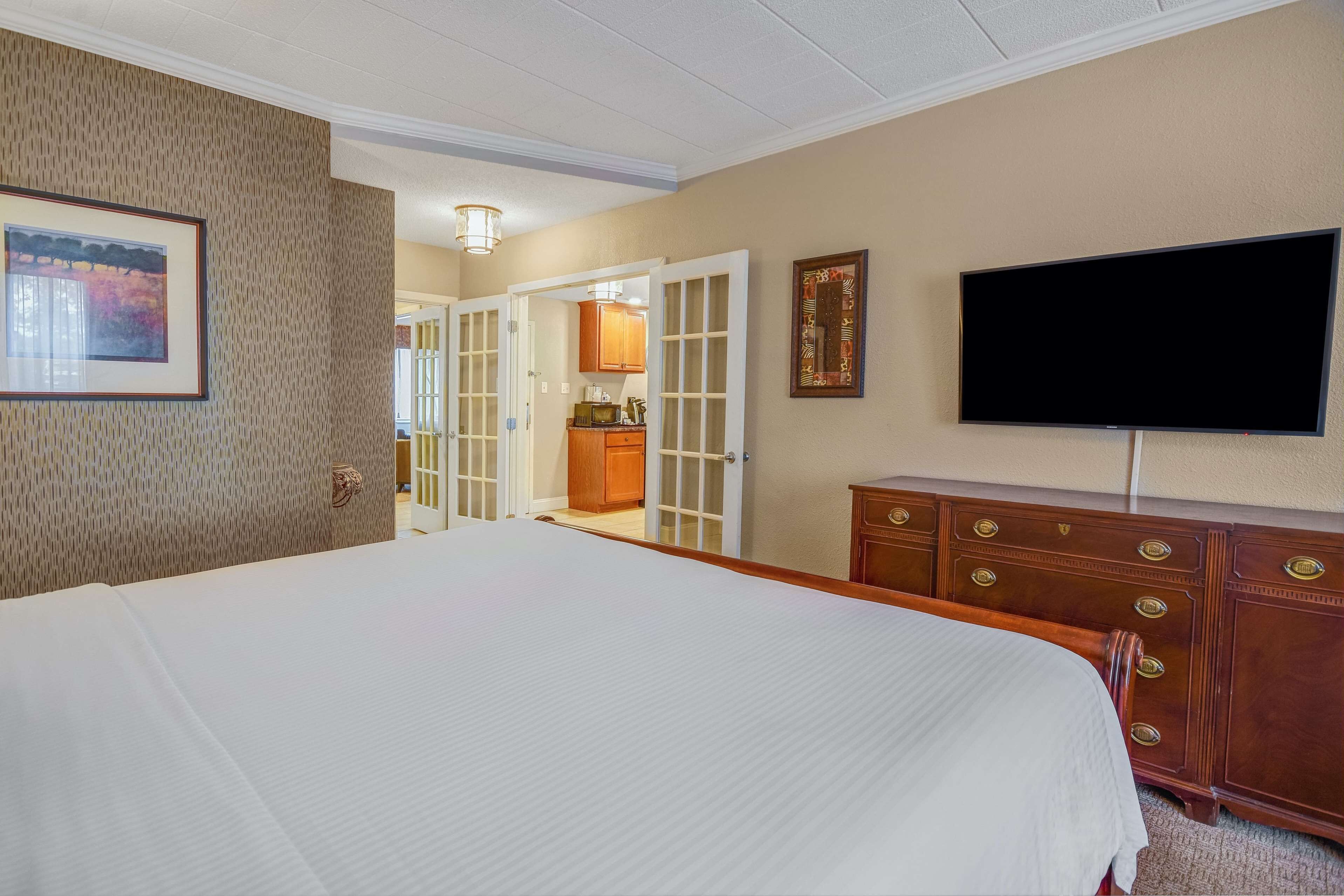 A hotel room with a large bed, a television, a wooden dresser, and a door leading to a kitchen area.