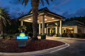 Een hotel met een receptiebord, een overdekte ingang en een verzorgd buitengebied met bomen en struiken.
