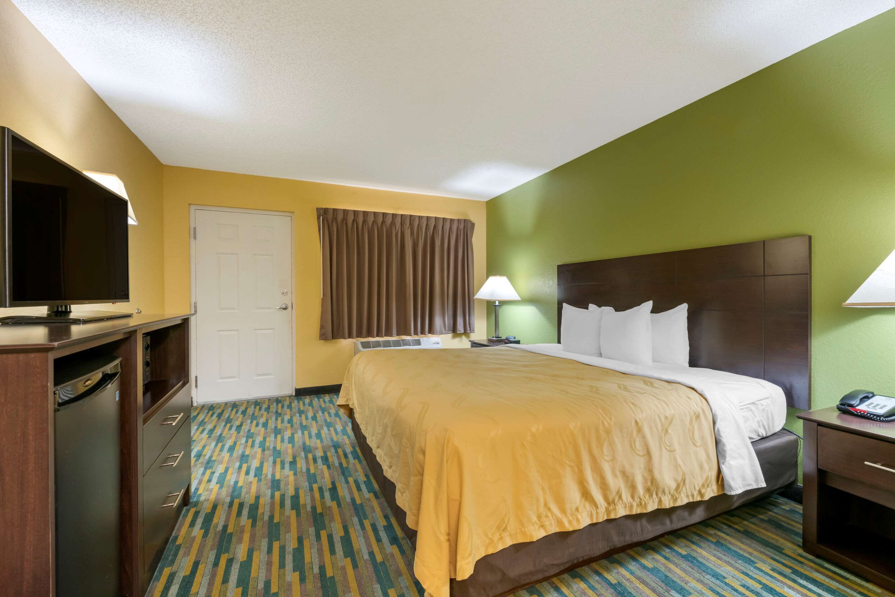 A hotel room with a bed, bedside table, lamp, and a television on a desk.