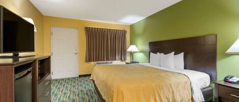 A hotel room with a bed, bedside table, lamp, and a television on a desk.