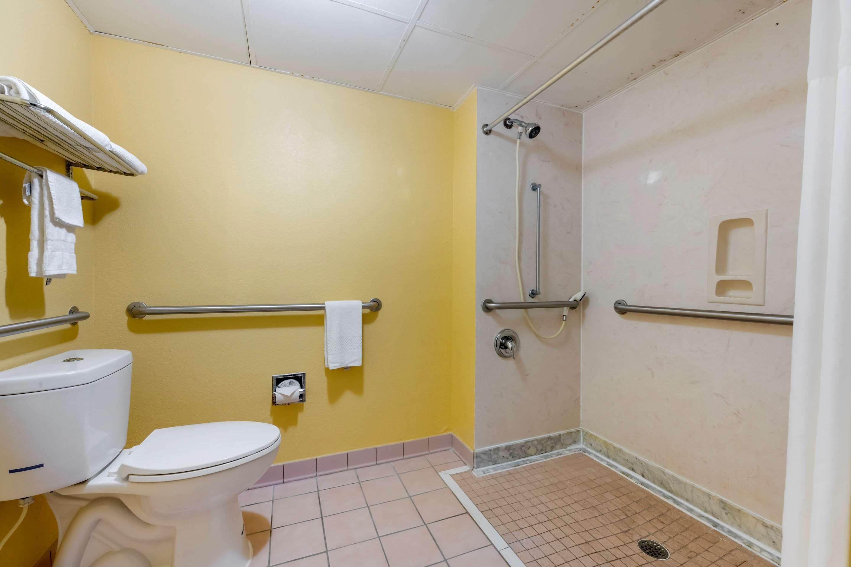 Un baño con pared amarilla, inodoro blanco, ducha con barras de apoyo y un perchero para toallas.