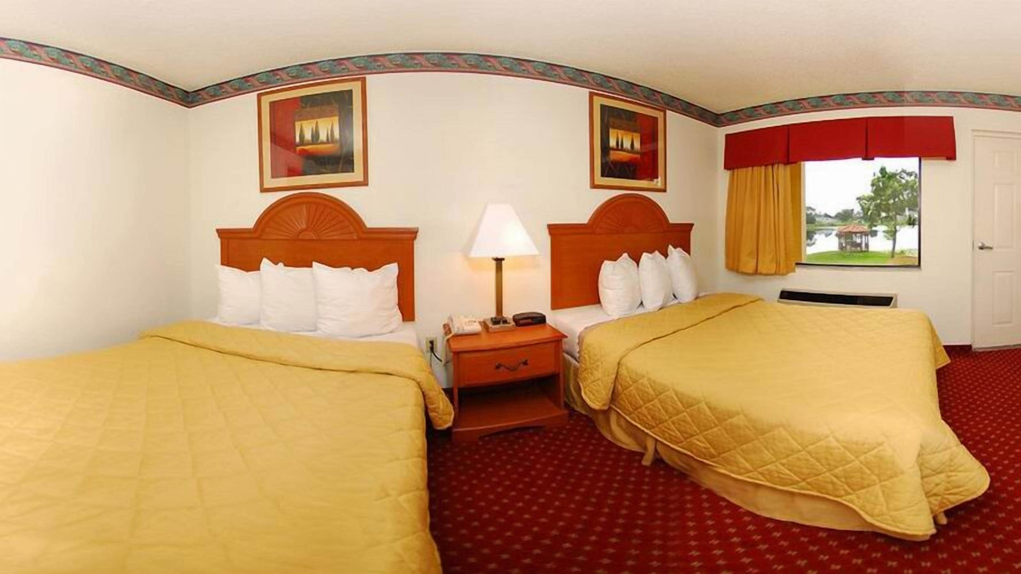 A hotel room with two beds, a nightstand, a lamp, and a window with a view of trees.