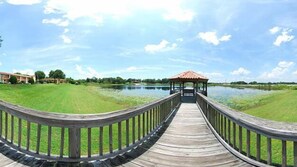 A wooden boardwalk leading to a gazebo over a body of water, surrounded by grassy areas and a building in the background.