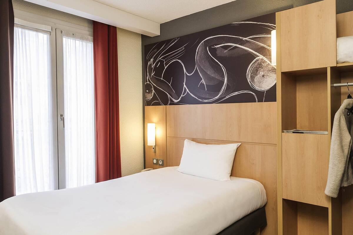 Habitación de hotel con una cama, una lámpara de pared, un armario de madera y una ventana con cortinas.