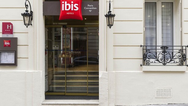 A hotel entrance with a red 'ibis HOTELS' sign, a glass door, and a balcony with wrought iron railings. - ibis Paris Vaugirard Porte de Versailles (Paris)