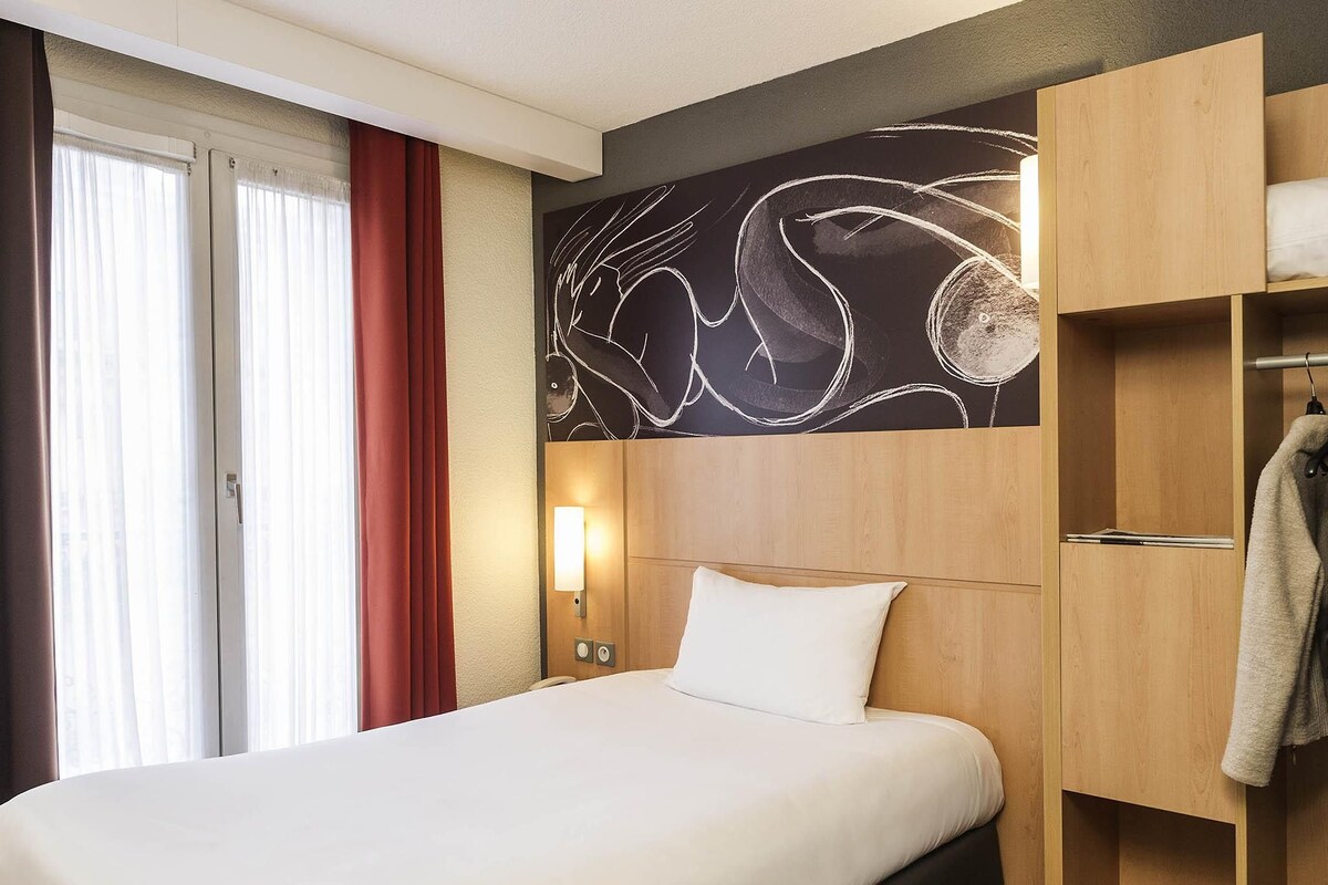 Ein Hotelzimmer mit einem Bett, einer Wandleuchte, einem Holzschrank und einem Fenster mit Vorhängen.