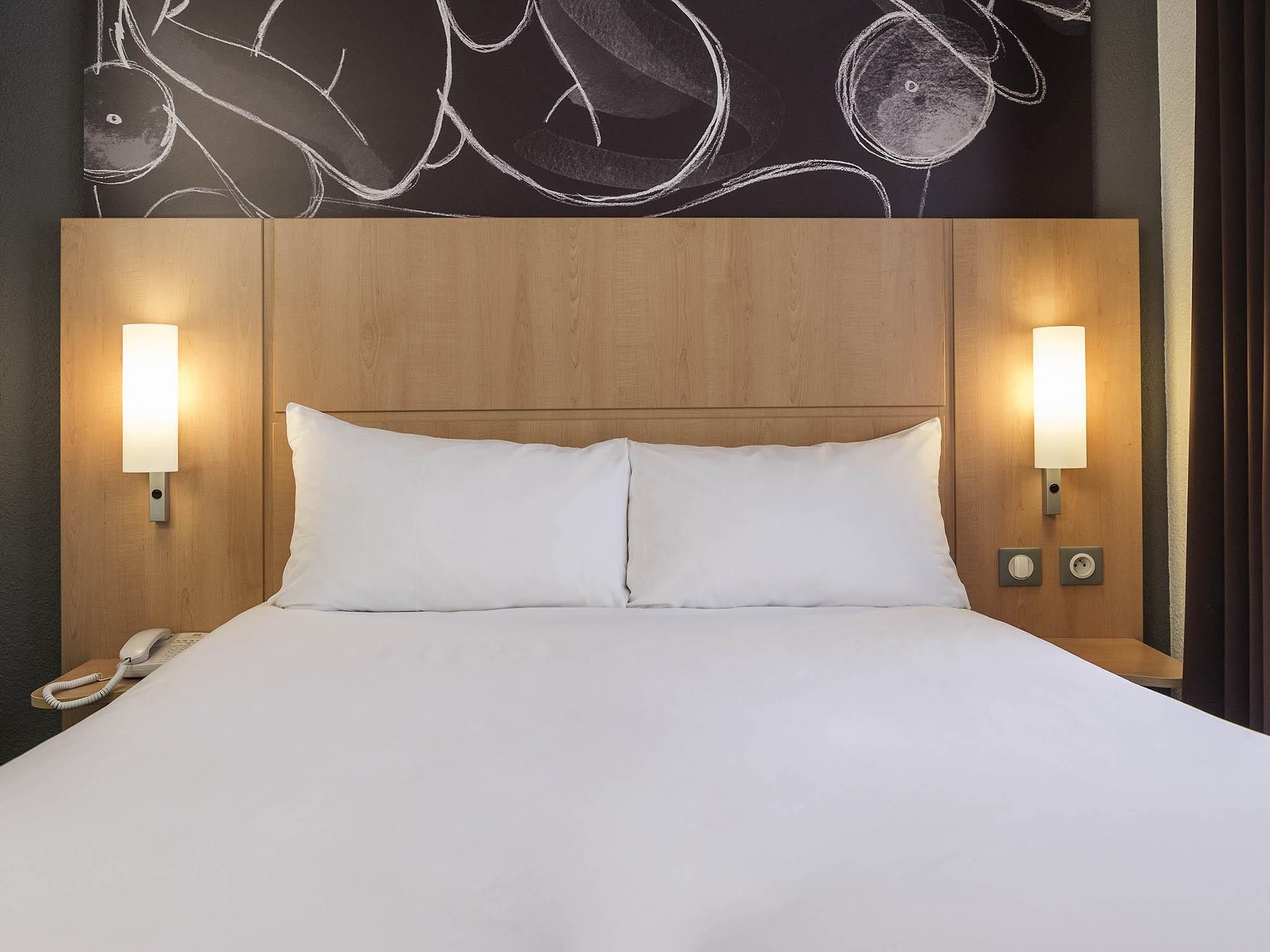 Une chambre d’hôtel avec une tête de lit en bois, deux lampes de chevet, un lit blanc et un téléphone sur la table de chevet.