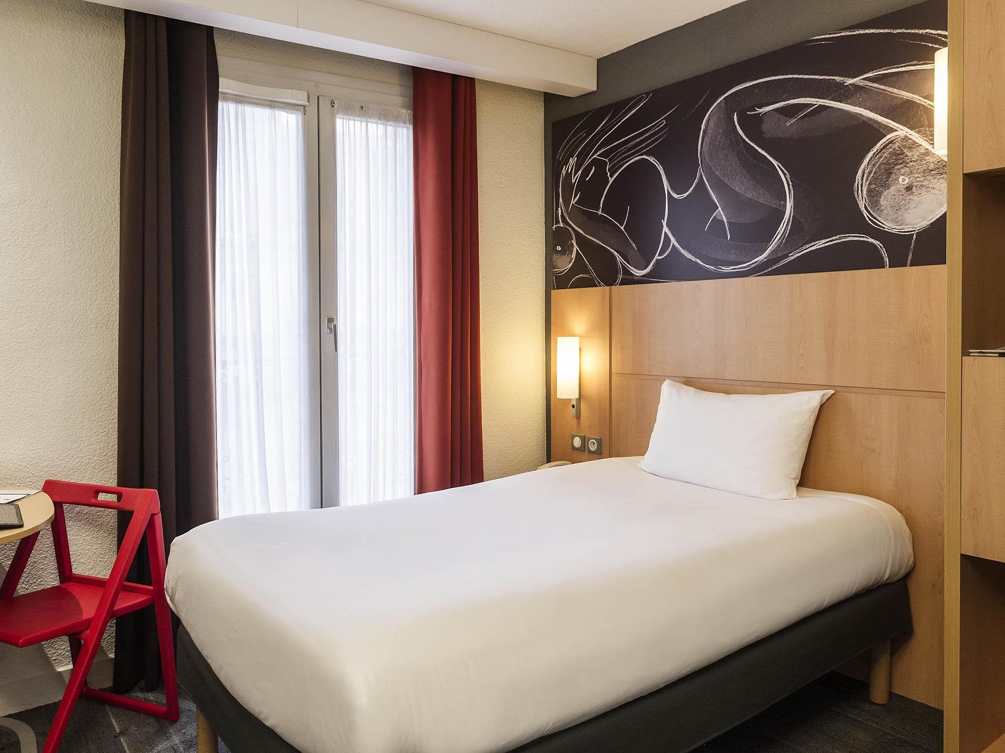 Une chambre d’hôtel avec un lit, un bureau, une chaise, une lampe fixée au mur et une fenêtre avec des rideaux.