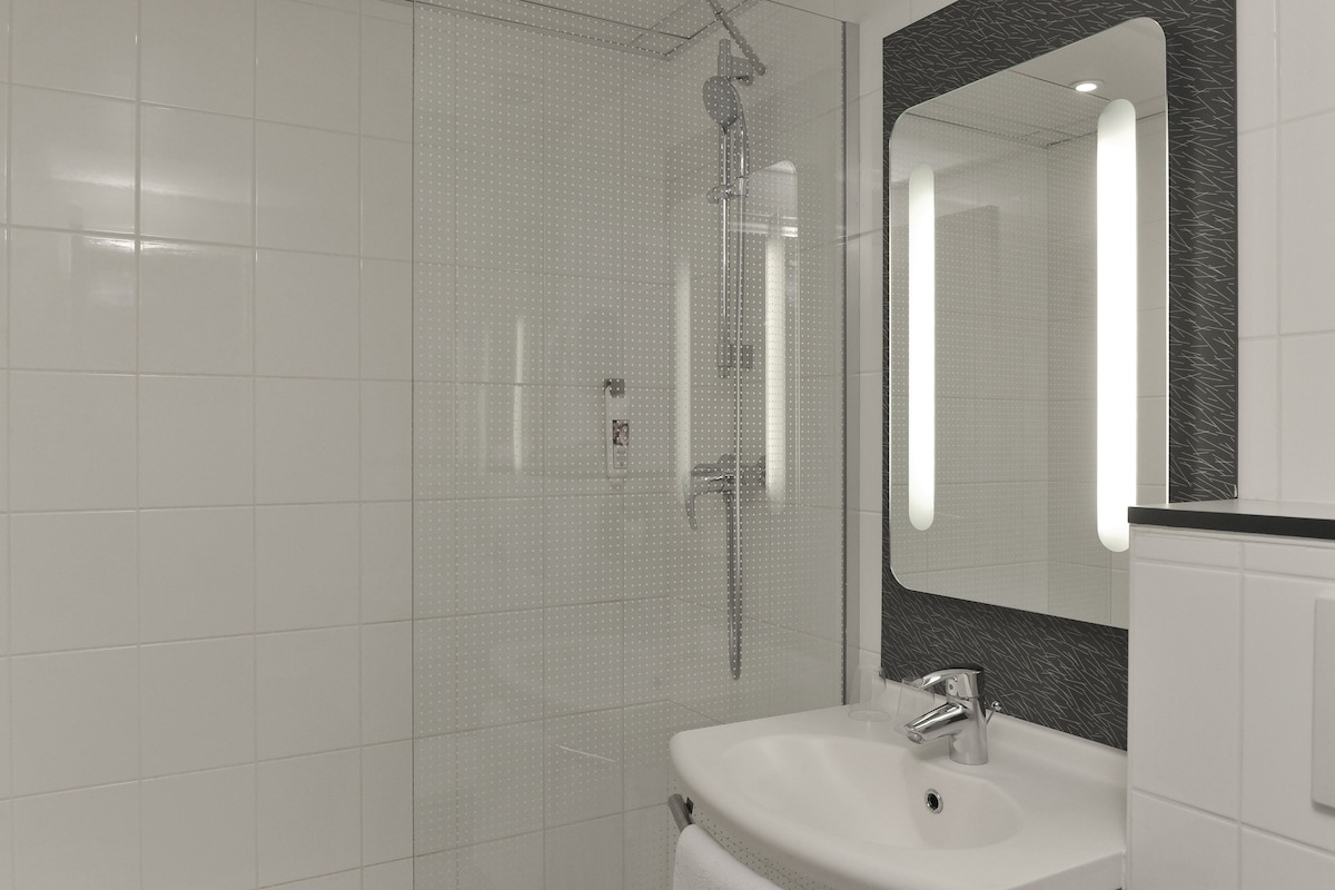 Une salle de bain moderne avec une douche en verre, un lavabo blanc et un grand miroir agrémenté d’un pare-splash foncé.