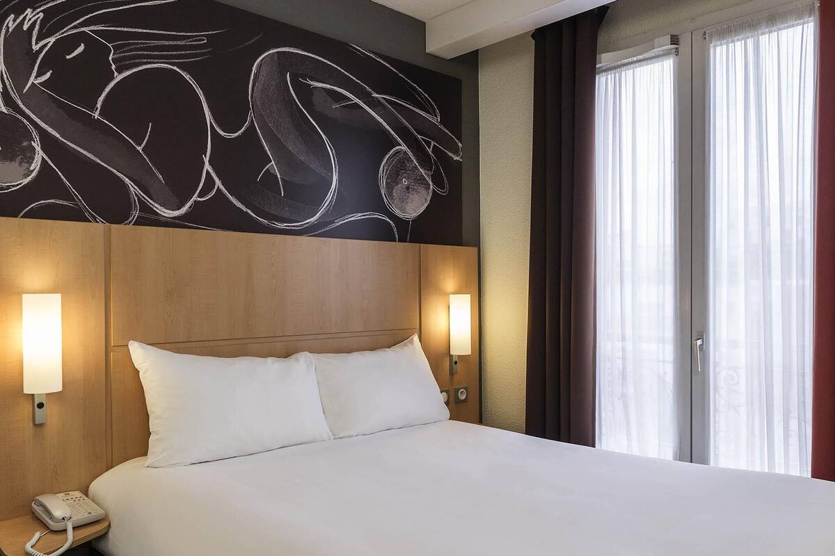 Habitación de hotel con una cama grande, cabecera con diseño artístico en la pared, lámparas de noche, teléfono en la mesita y una ventana con cortinas.