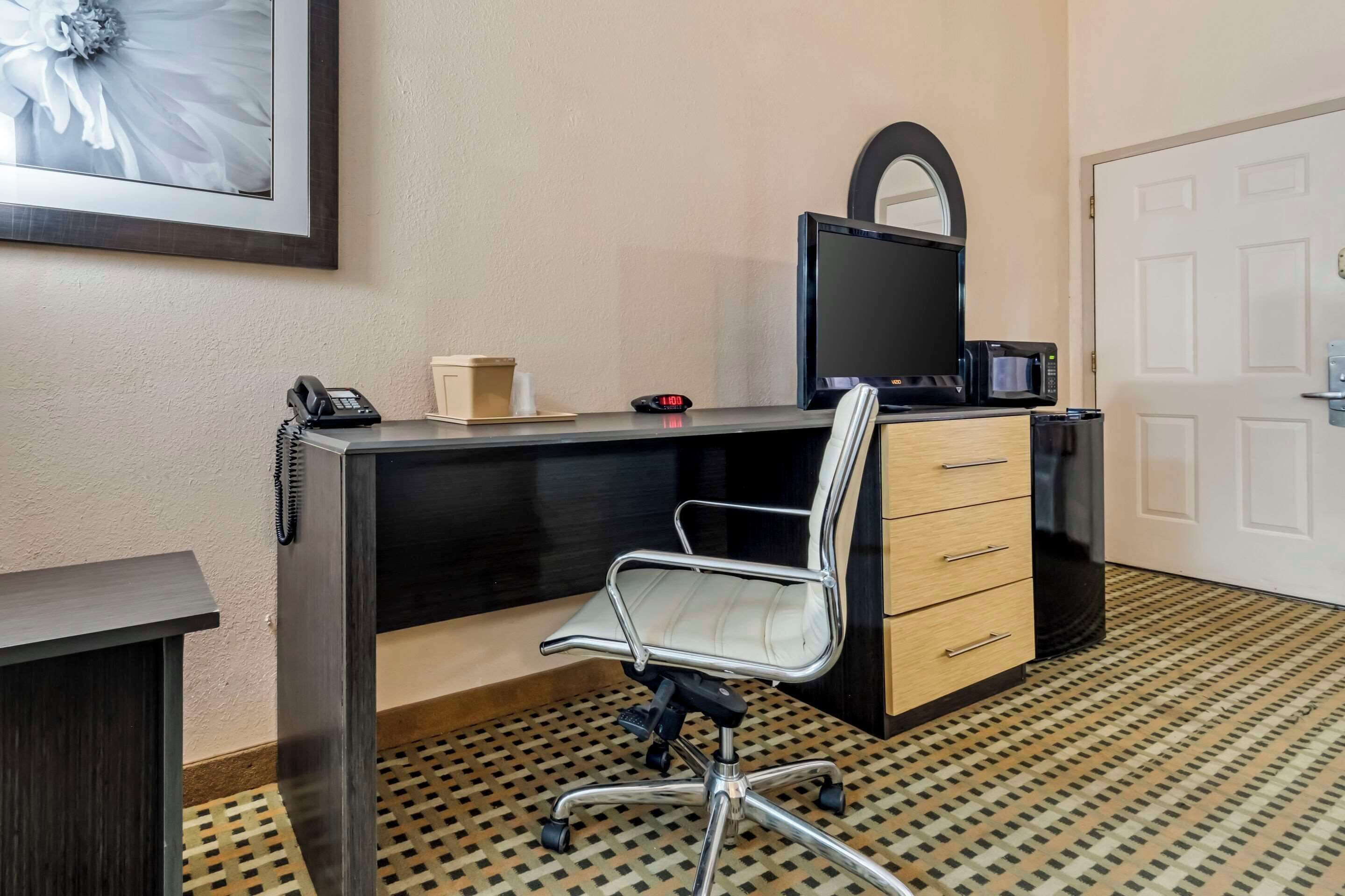 A hotel room with a desk, a chair, a telephone, a clock, a television, and a microwave.
