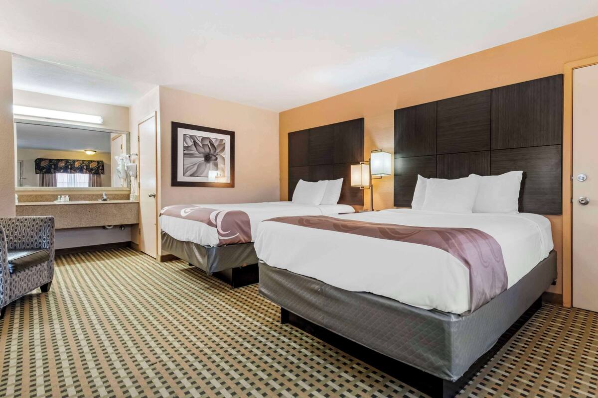 Una habitación de hotel con dos camas, un espejo grande, una obra de arte enmarco, un escritorio y una alfombra estampada.