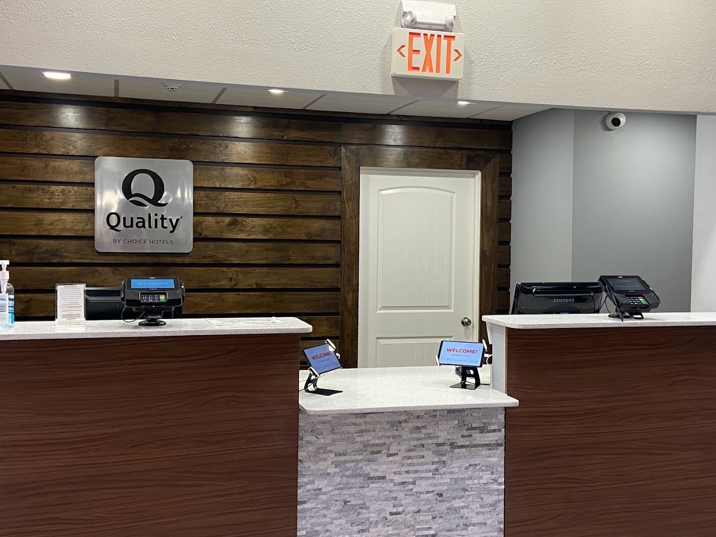 A hotel reception desk with a 'Quality by Choice Hotels' sign, an 'EXIT' sign above, and a 'WELCOME!' sign below.