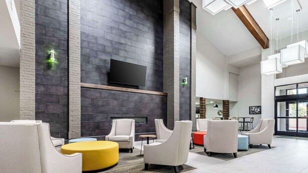 A modern lobby with a flat-screen TV mounted on a dark brick wall, surrounded by white armchairs and a yellow ottoman.