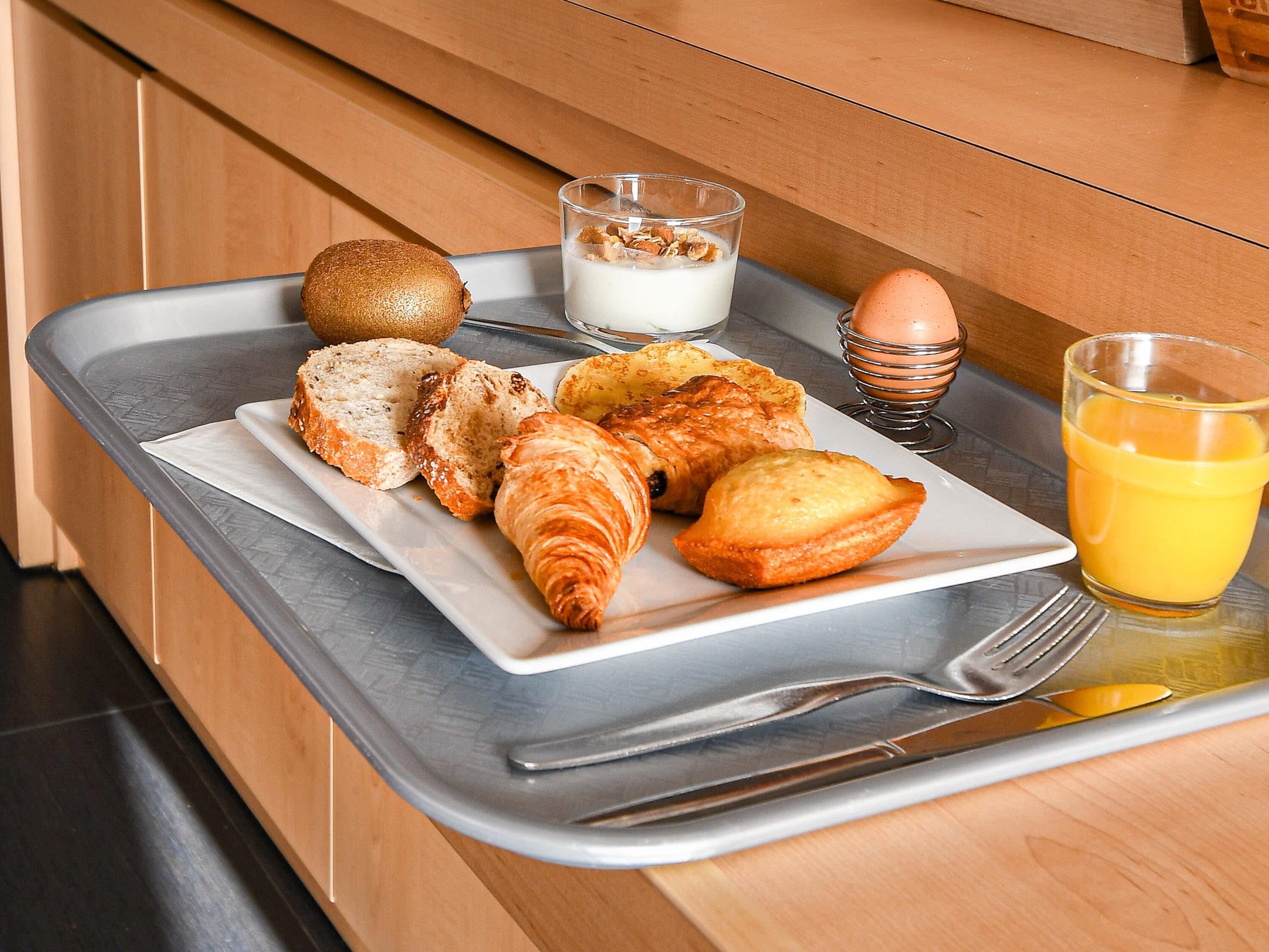 A breakfast tray with croissants, bread, a kiwi, a glass of orange juice, a glass of milk with granola, and a boiled egg on a kitchen counter.