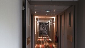 Un pasillo de hotel con alfombra a rayas, puertas a ambos lados y un extintor en la pared derecha.
