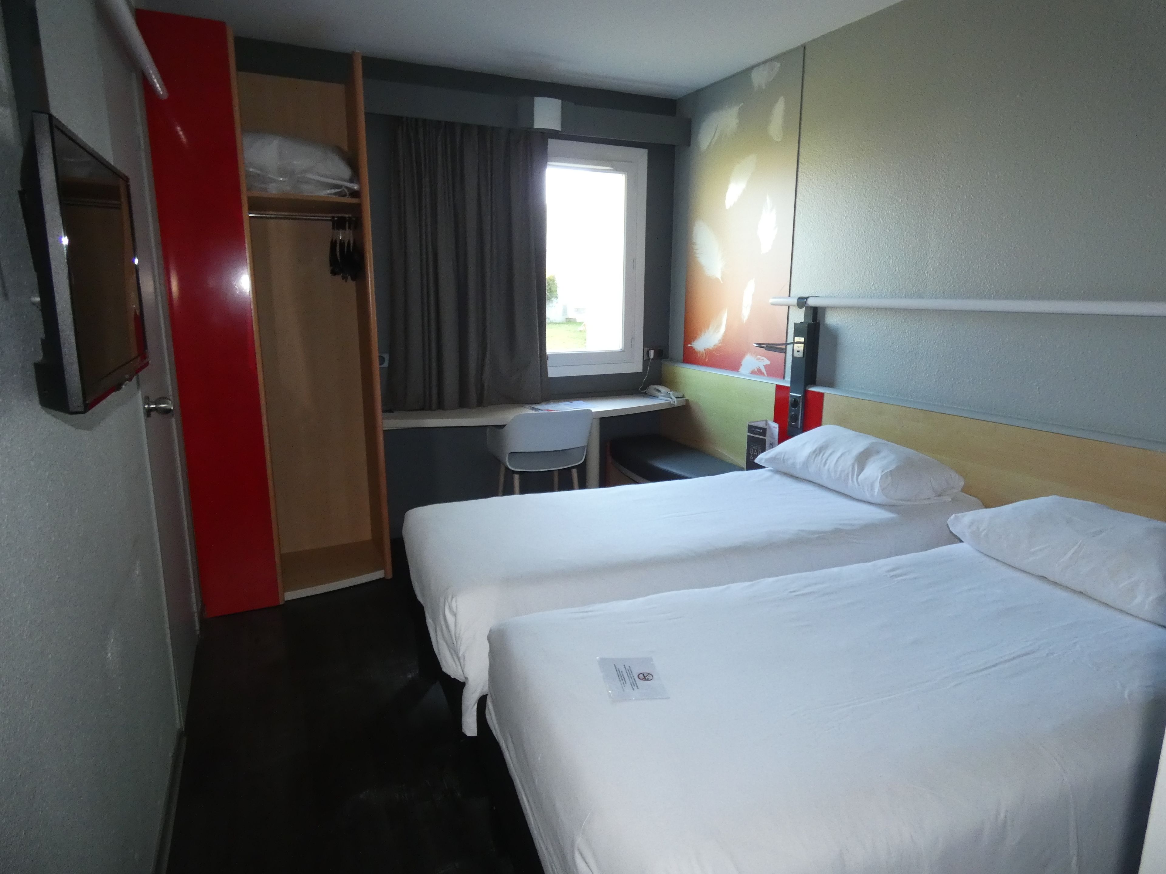 Quarto de hotel com duas camas, uma escrivaninha, uma cadeira e uma janela com cortinas.