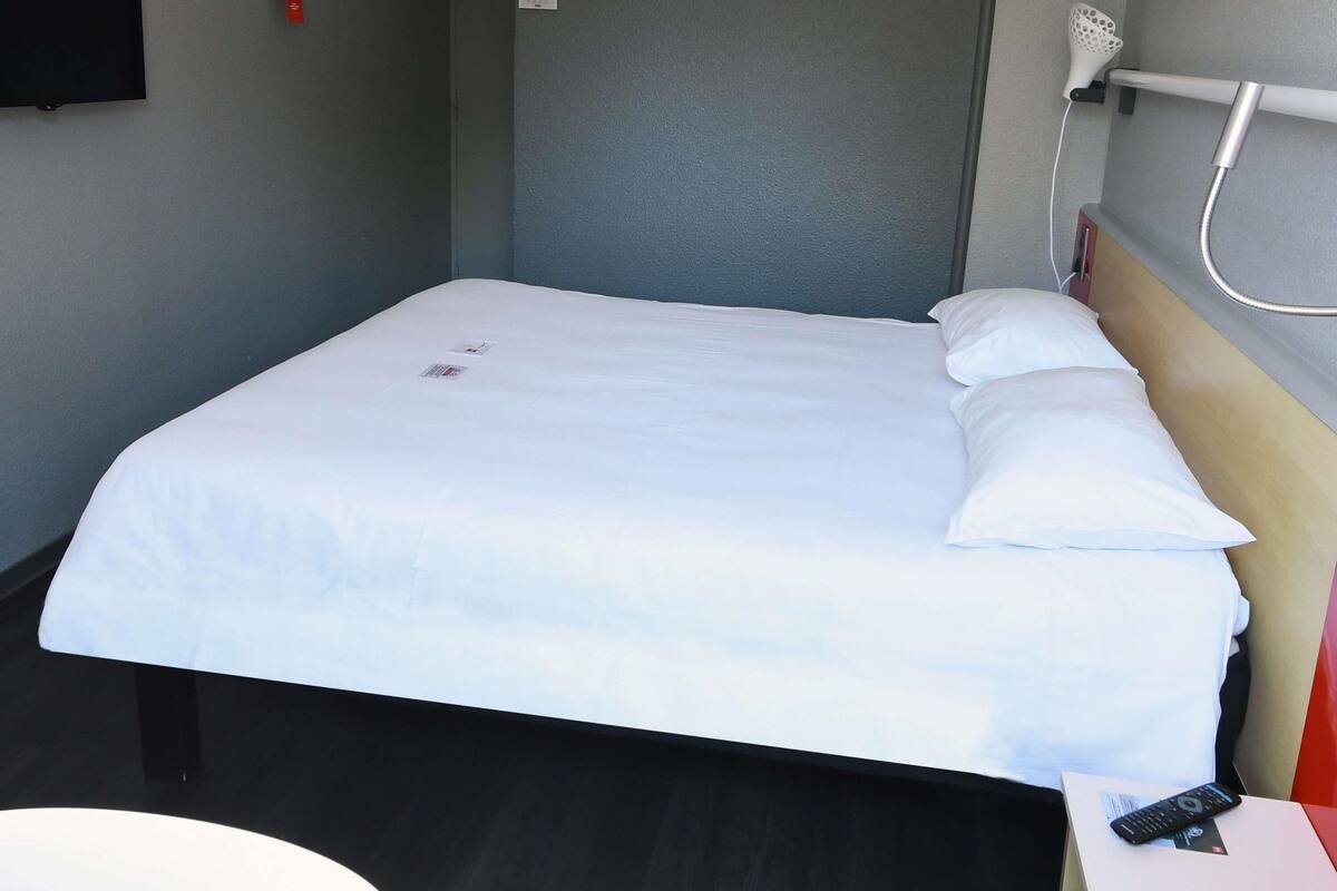 Ein Einzelbett mit weißer Bettwäsche, eine Fernbedienung auf einem kleinen Tisch und ein Duschkopf an der Wand.