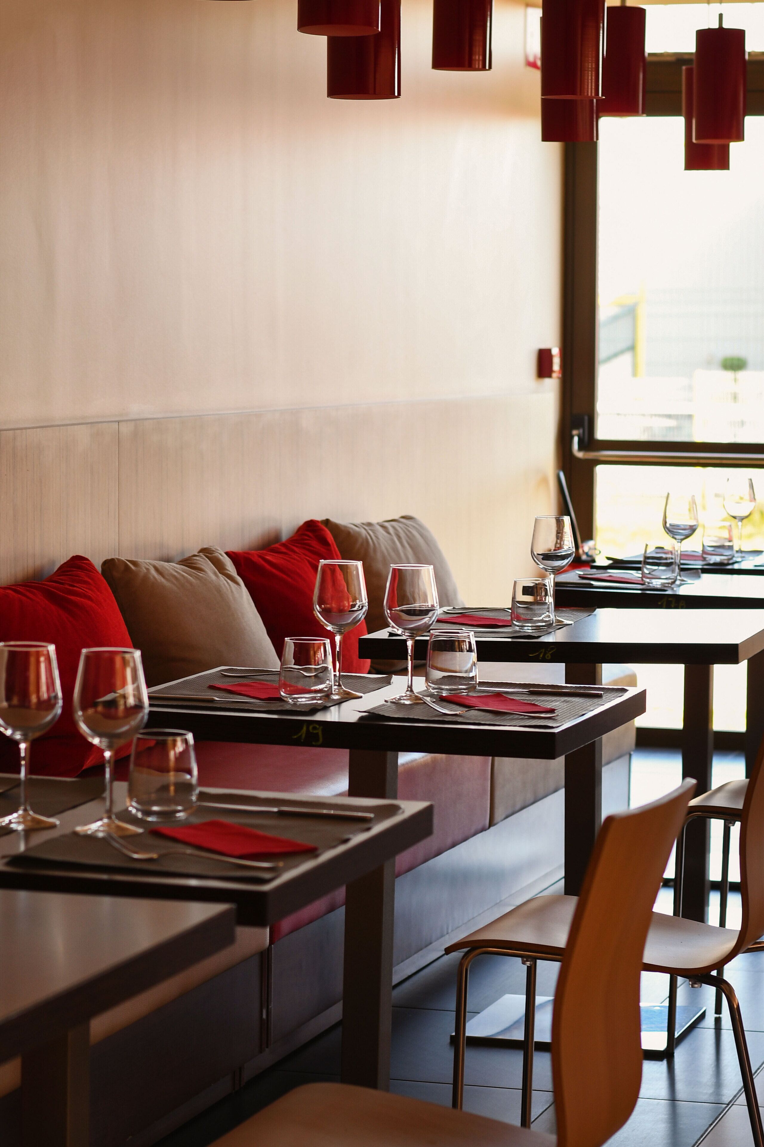 A restaurant with tables set for dining, red cushions on the seating, and hanging red pendant lights.