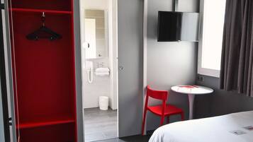 Una camera d'albergo con un letto, un armadio rosso, un piccolo tavolo con una sedia, una TV montata a parete e un bagno visibile attraverso una porta aperta.