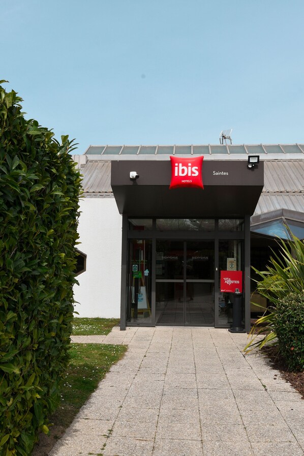 The exterior of an ibis hotel with a modern design, featuring a prominent red and white logo, glass doors, and a well-maintained entrance area.