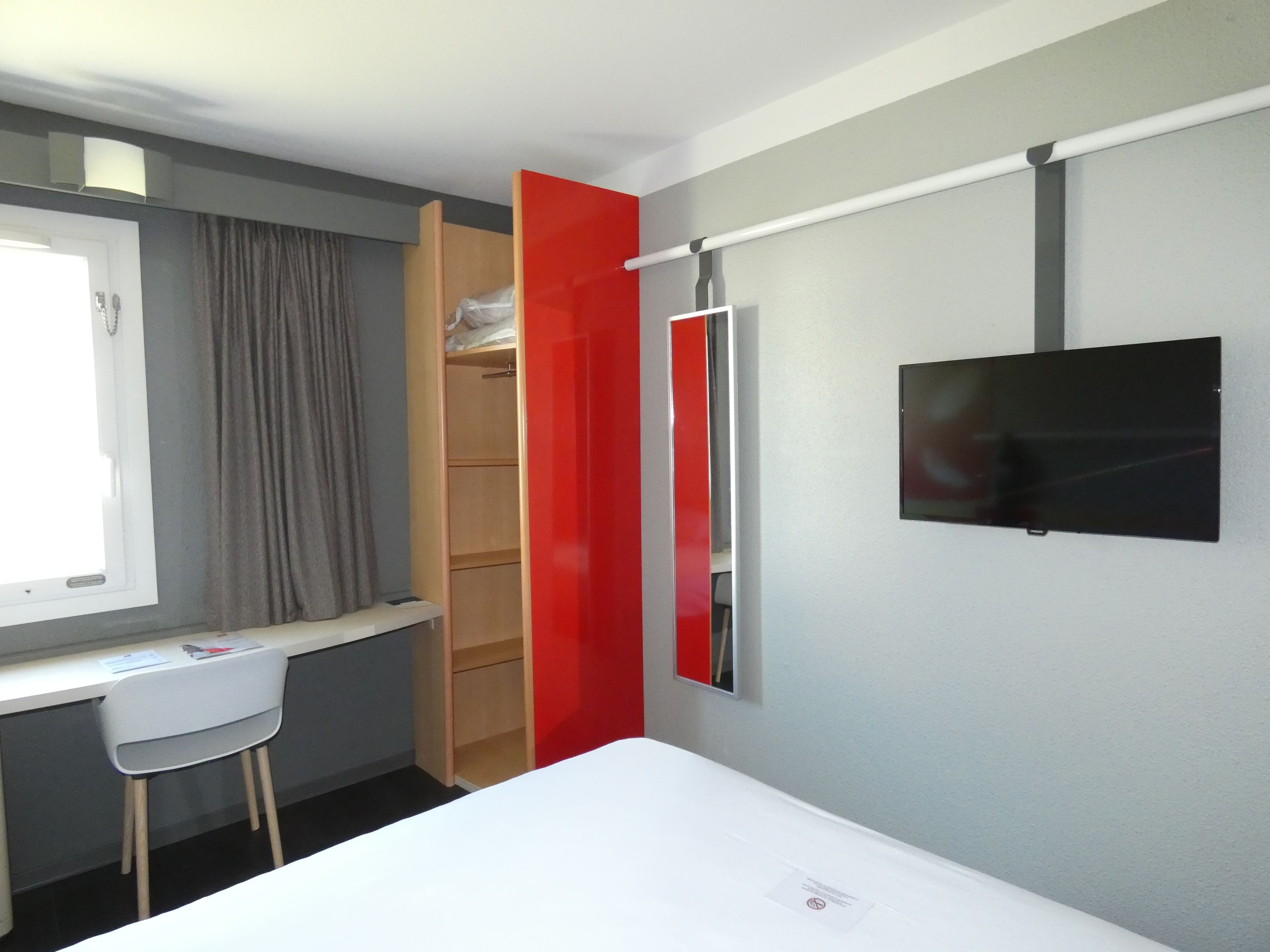 Habitación de hotel pequeña con cama, escritorio, silla, armario y televisión montada en la pared.