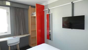 Habitación de hotel pequeña con cama, escritorio, silla, armario y televisión montada en la pared.