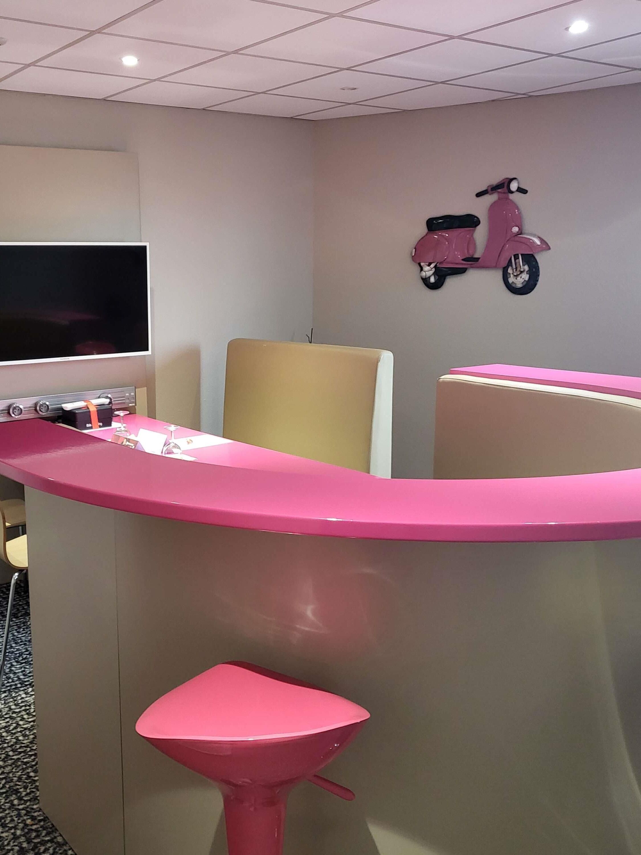 Een receptiegedeelte met een roze balie, een televisie, een scooterdecoratie aan de muur, en een enkele stoel.