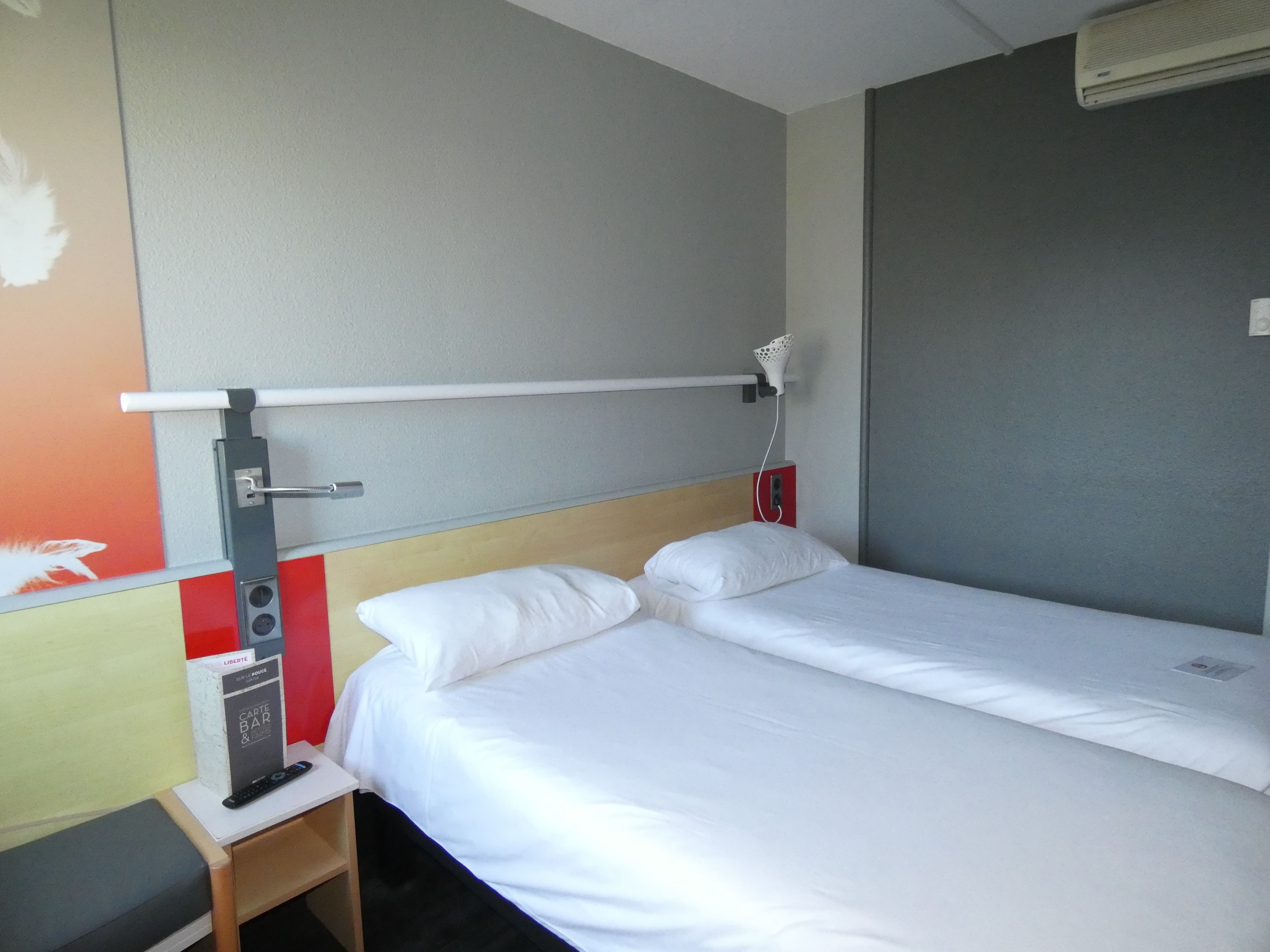 Une chambre d’hôtel avec un lit simple, une petite table de chevet avec une télécommande, un climatiseur fixé au mur et un mur rouge orné d’une décoration en forme de plume blanche.