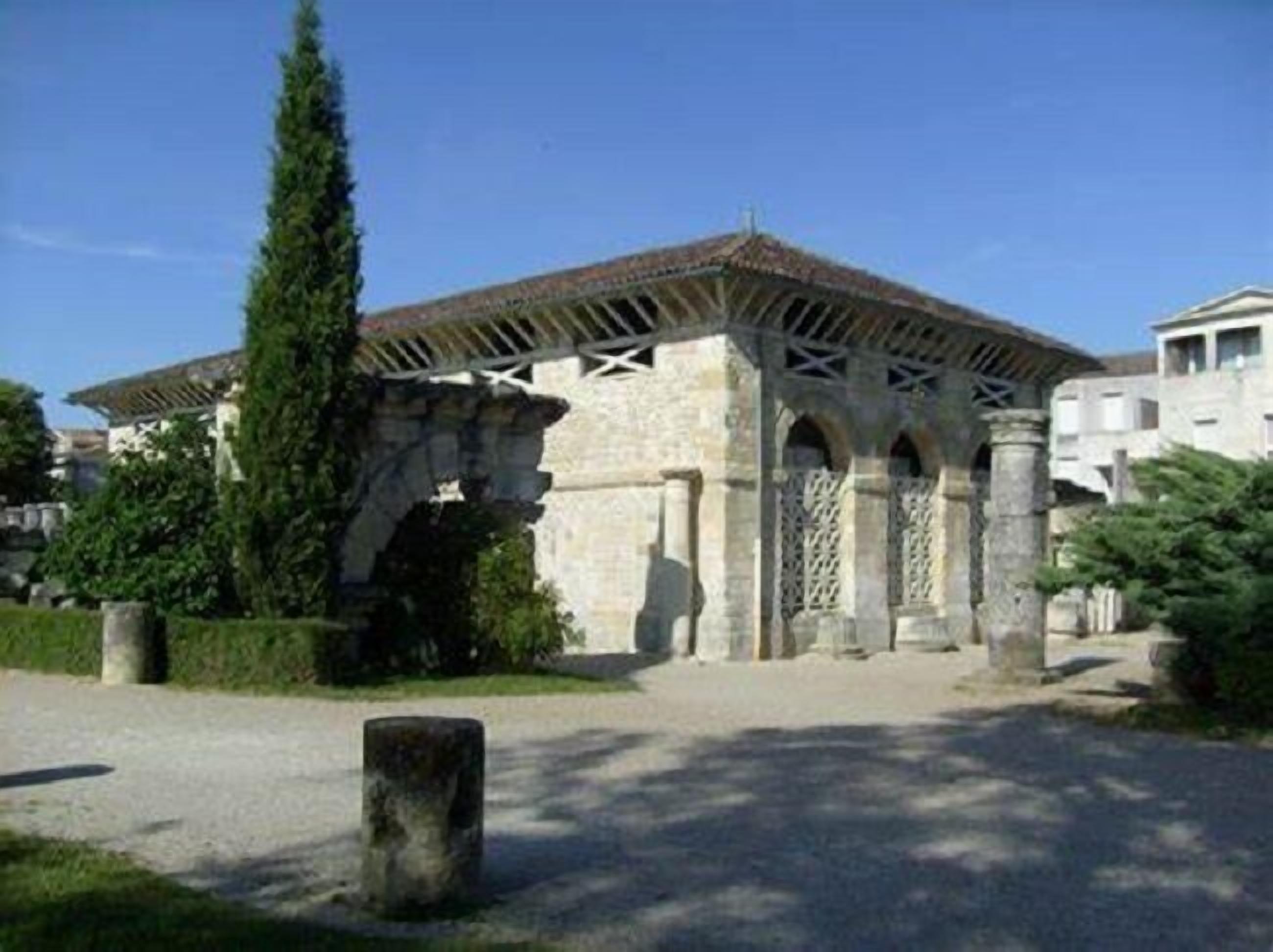 A historical stone building with a tiled roof and arched openings, surrounded by greenery and modern buildings.
