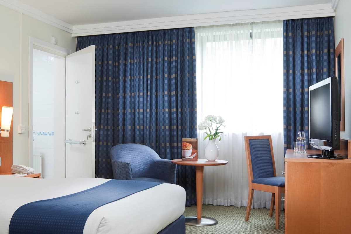 A hotel room with a large bed, a desk with a chair, a television, and a small table with a vase of flowers.