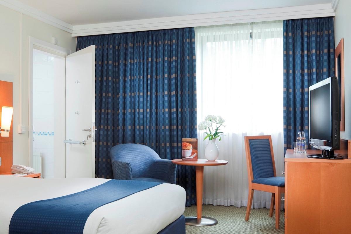 A hotel room with a bed, a desk with a chair, a television, and a window with curtains.