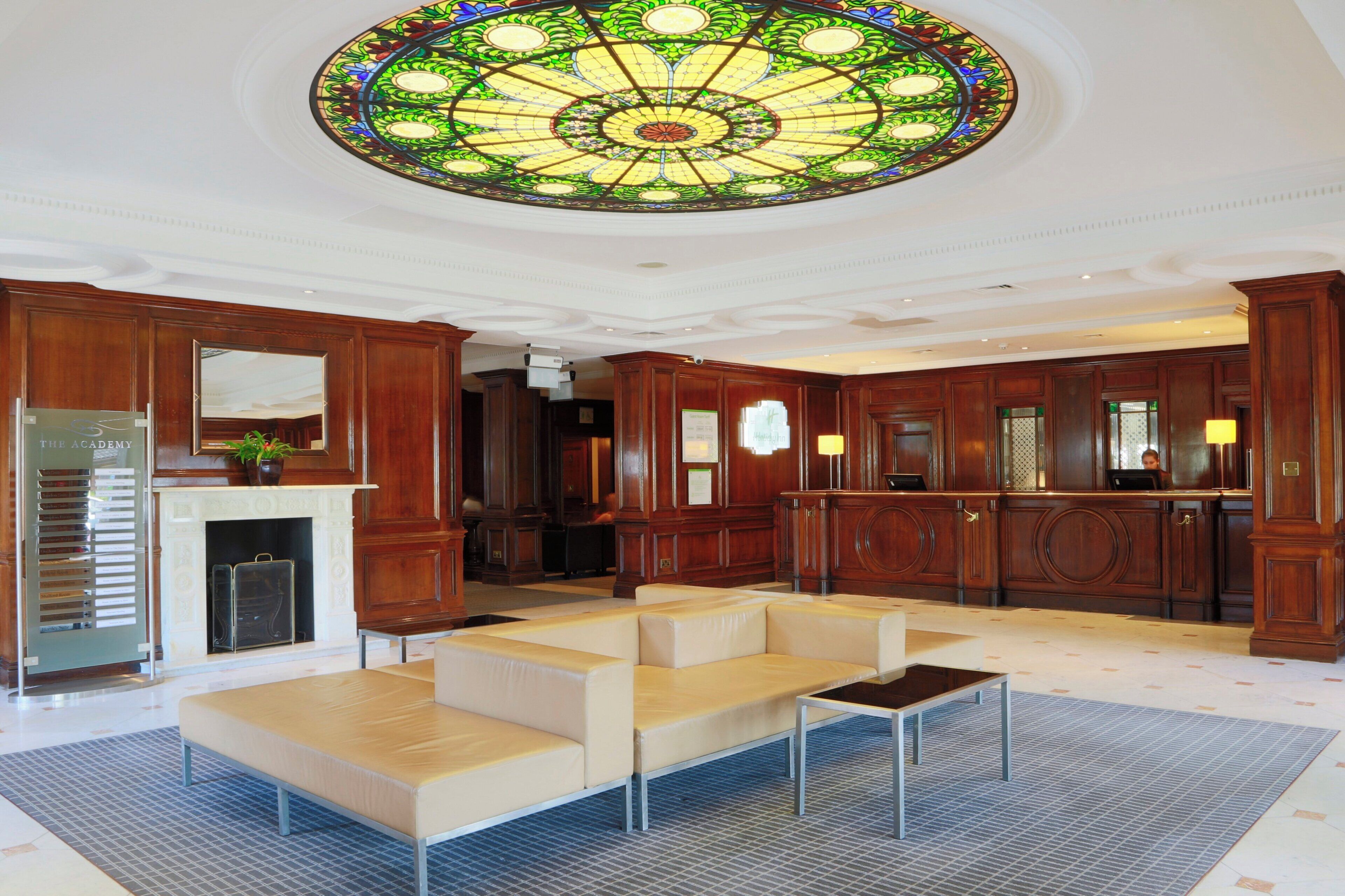 A hotel lobby with a large stained glass ceiling, wooden paneling, a fireplace, and a seating area.