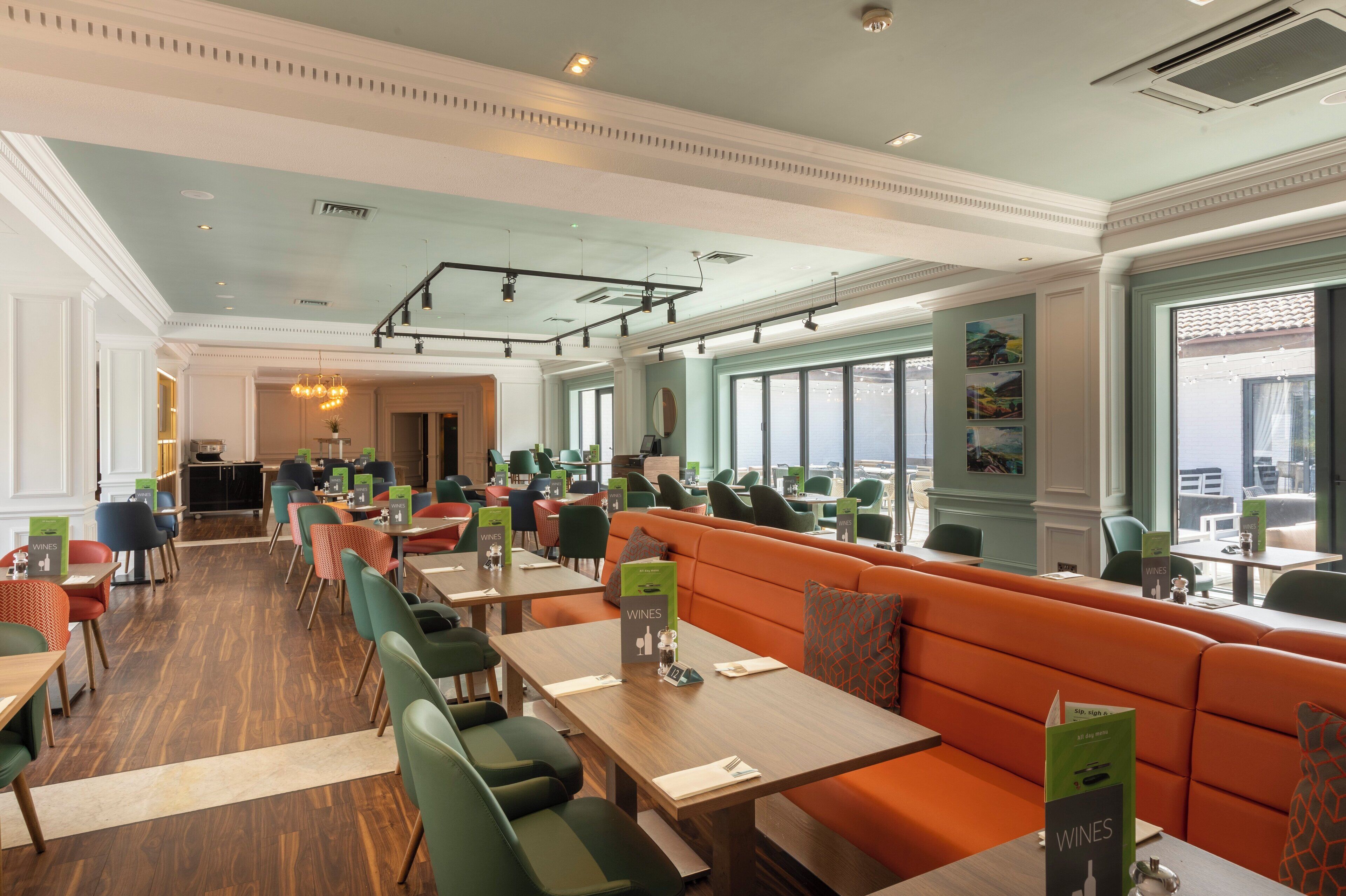 Un restaurante espacioso con techos altos, ventanas grandes y diversas opciones de asientos, incluyendo mesas y un amplio banco naranja.