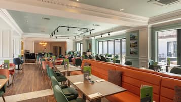 Un restaurante espacioso con techos altos, ventanas grandes y diversas opciones de asientos, incluyendo mesas y un amplio banco naranja.