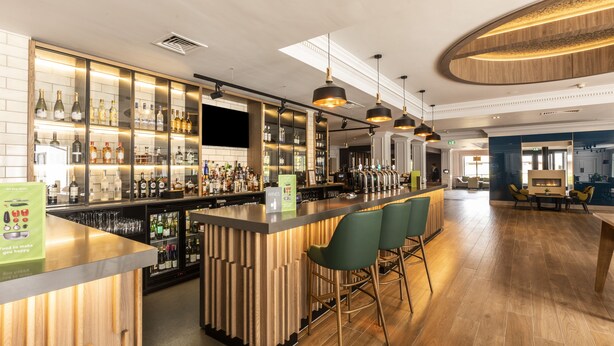 A bar area with wooden bar counters, green bar stools, and a variety of bottles and glasses. - Holiday Inn Guildford by IHG (Guildford)