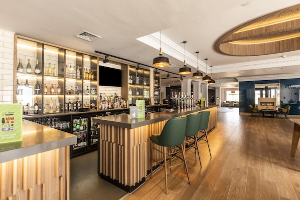 A bar area with wooden bar counters, green bar stools, and a variety of bottles and glasses.