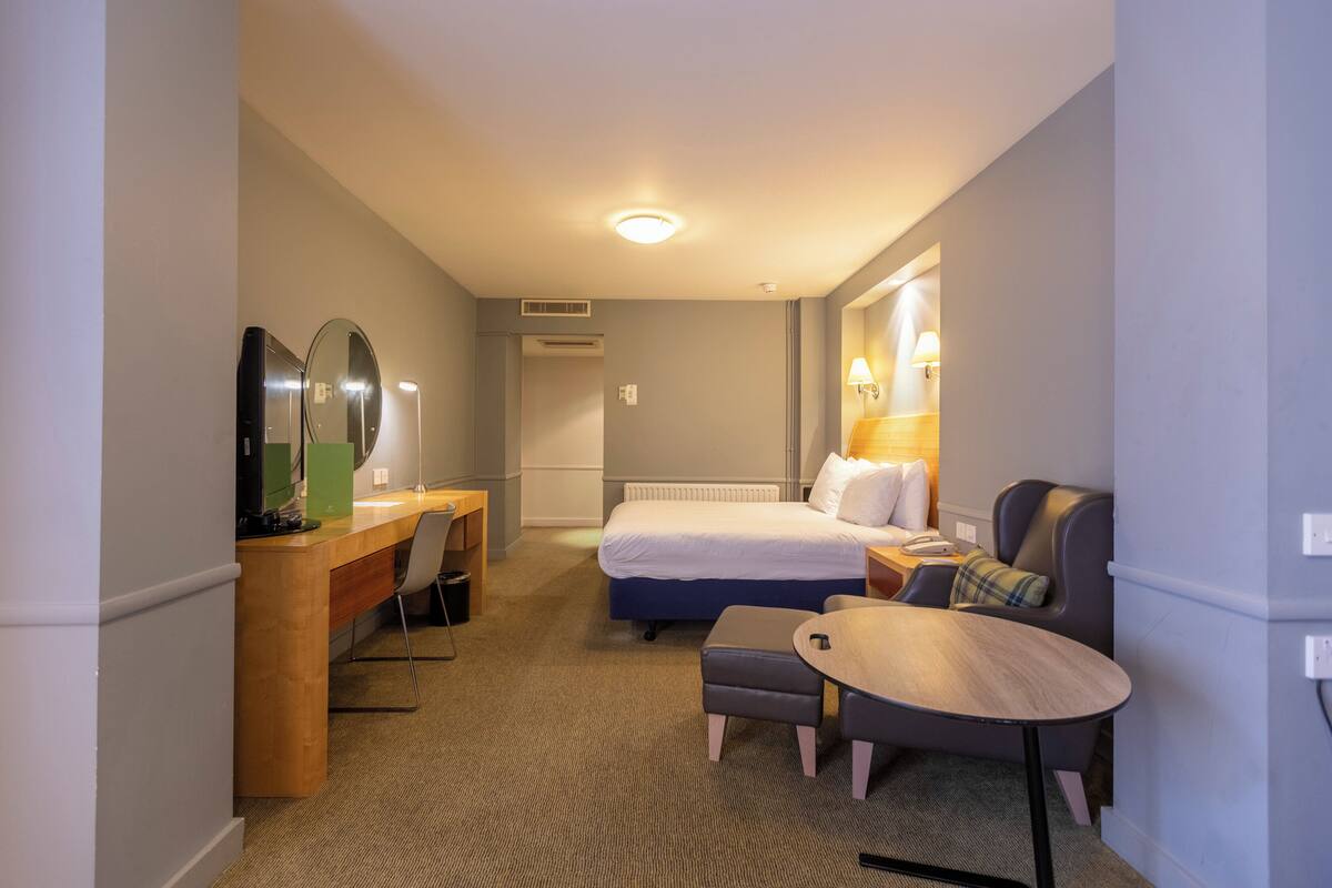 A hotel room with a bed, a desk, a chair, a small table, and a lamp.