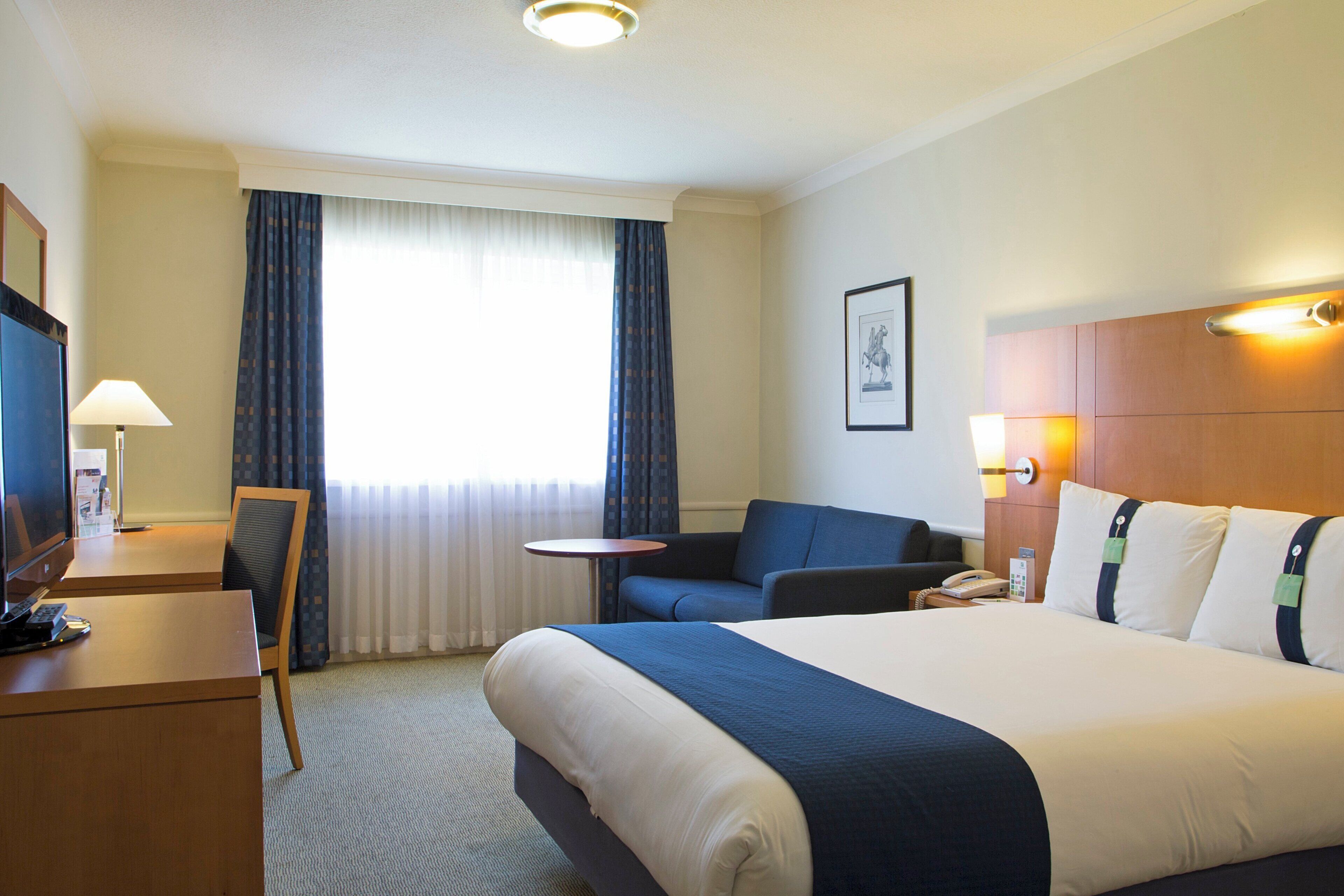A hotel room with a bed, a desk, a chair, a small table, and a window with curtains.