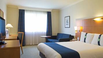 A hotel room with a bed, a desk, a chair, a small table, and a window with curtains.