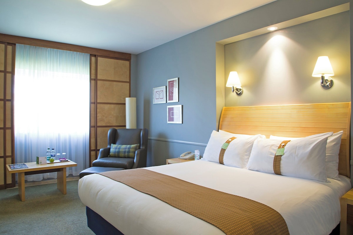 A hotel room with a large bed, a desk, a chair, and two wall-mounted lamps.