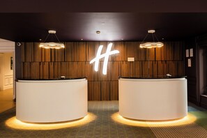 A reception area with two white reception desks, a wooden wall with a large illuminated 'H', and pendant lighting.