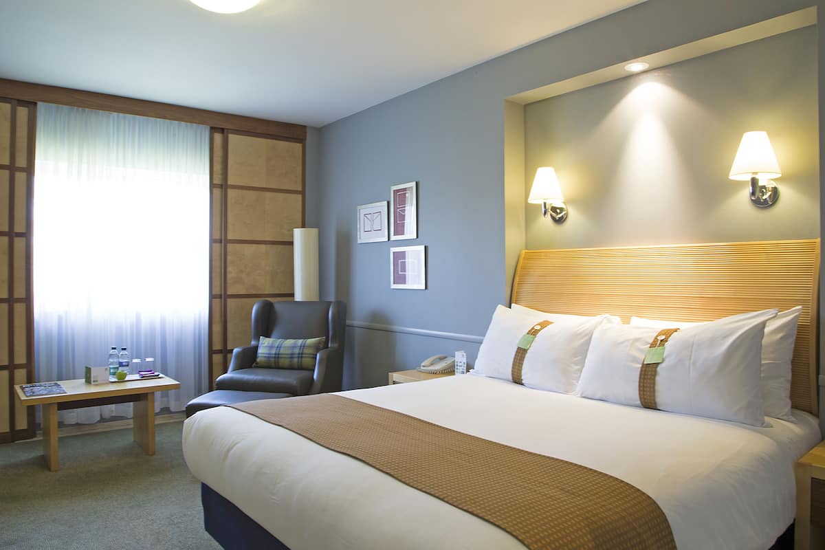 A hotel room with a large bed, a desk, a chair, and two wall-mounted lamps.