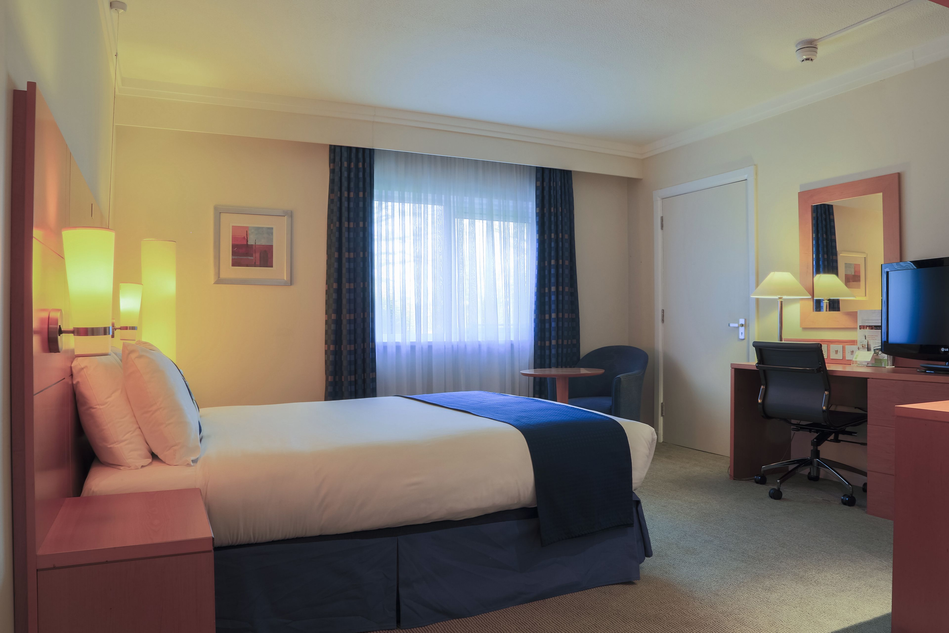 A hotel room with a bed, bedside lamps, a desk with a chair, a television, and a window with curtains.