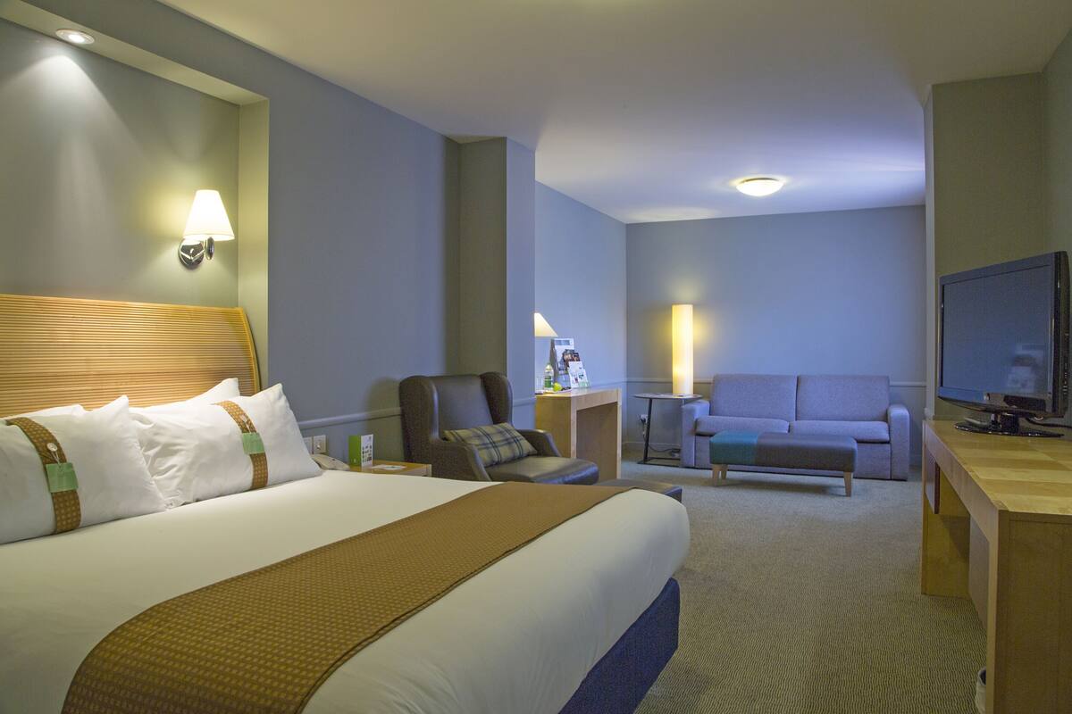 A hotel room with a large bed, a desk, a television, a sofa, and a small table with a lamp.