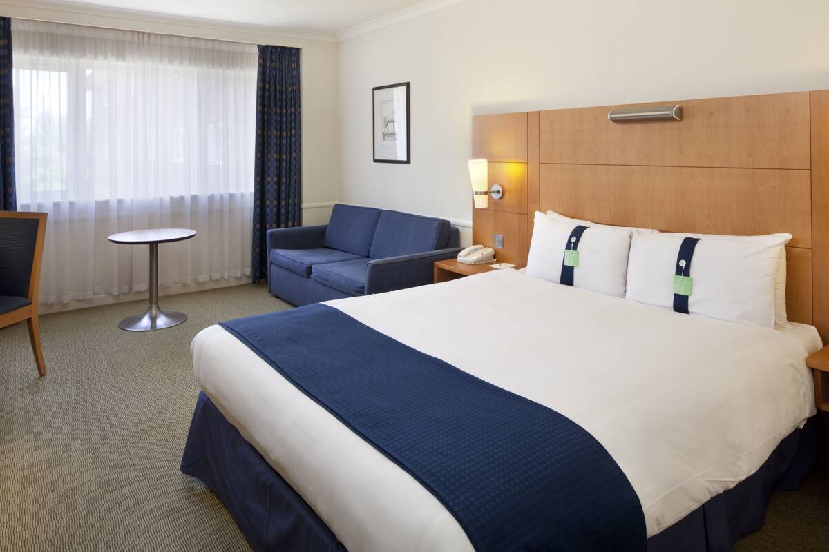 A hotel room with a large bed, a small round table, a sofa, and a nightstand with a lamp.