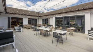An outdoor patio area with multiple tables and chairs, a wooden deck, and a building with large windows and a tiled roof.