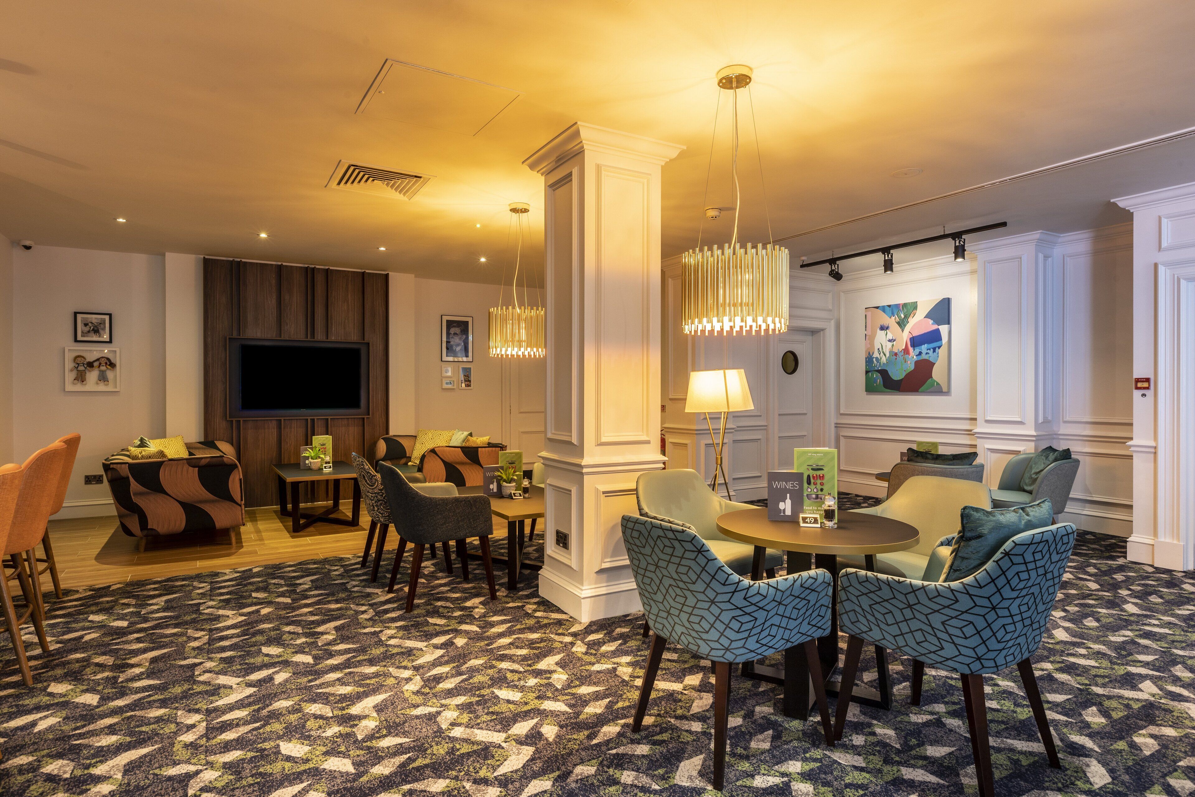 Un hall de hotel con una televisión de pantalla plana grande, una zona de estar con sillas estampadas, una mesa de centro y una lámpara decorativa.
